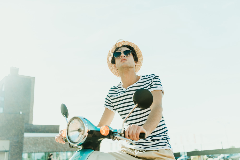 Millennial on scooter wearing hat and sunglasses looking up towards the blue sky