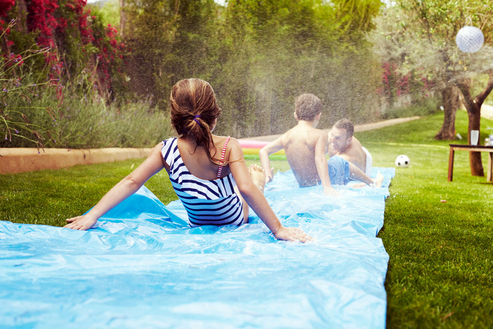 family slides down blue plastic slip-n-slide on beautiful green lawn
